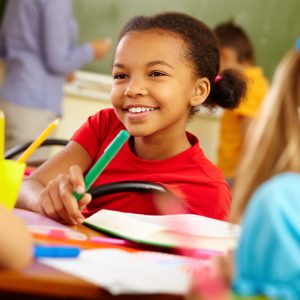 Portrait of cute girl holding crayon while looking at classmate at lesson